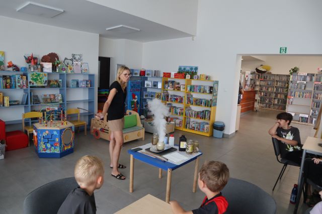 edukatorka stoi przed stolikiem na którym wytworzyła dym, w tle regały