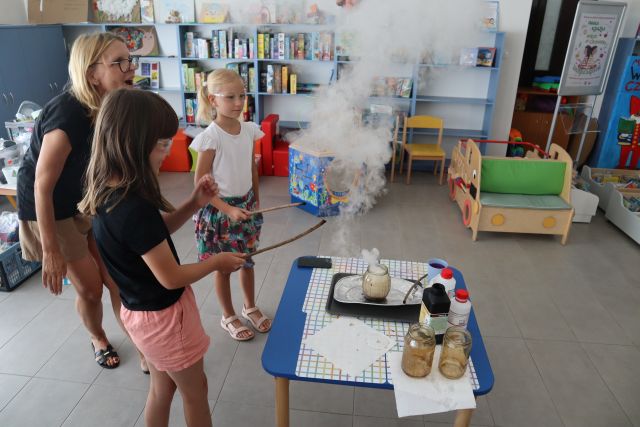 dwie dziewczynki przy pomocy kobiety patykiem czarują dym