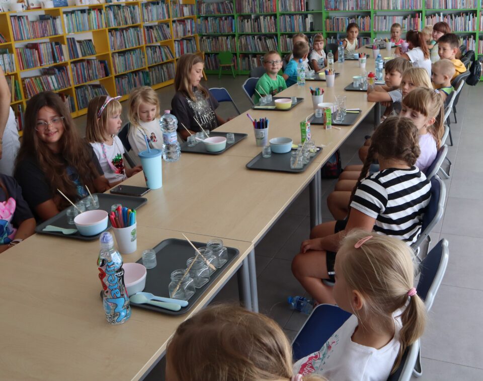 30 dzieci siedzi przy połączonych w rząd stołach, przed nimi na stołąch tacki, słoiczki i miseczki, w tle regały z książkami