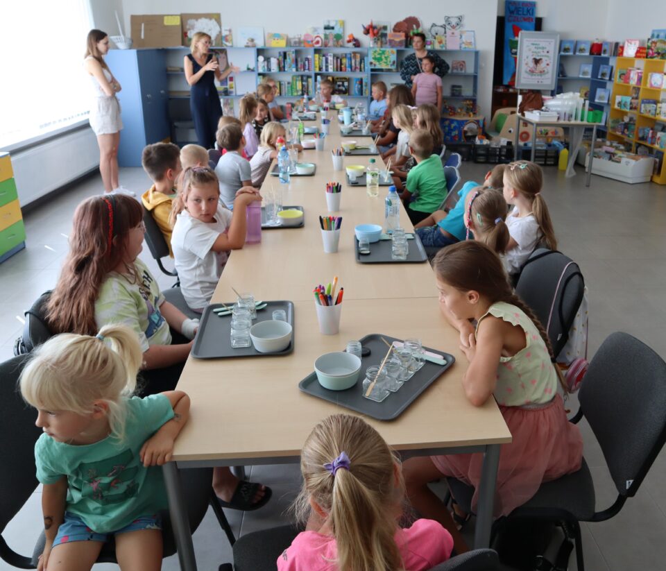 30 dzieci siedzi przy stołach, za nimi stoją 3 kobiety, w tle regały z książkami