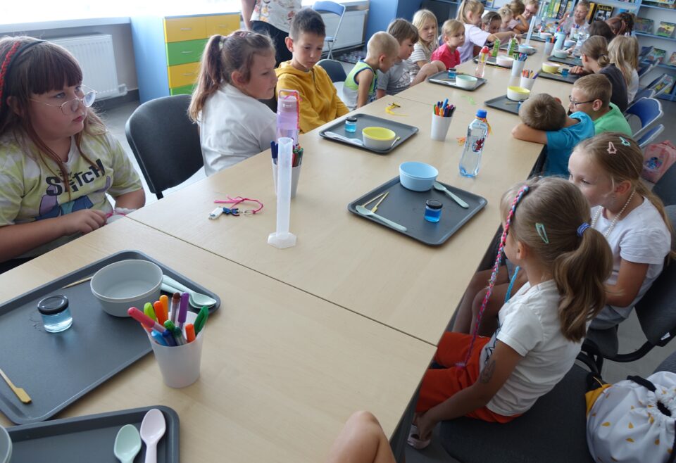 dzieci siedzące przy stołach obserwują kolorową ciecz w słoiczkach