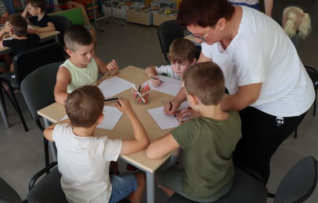 kobieta pomaga dzieciom siedzącym przy stoliku narysować komiks