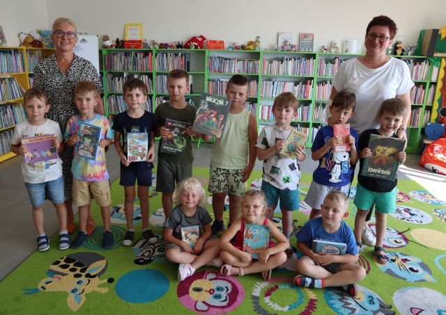 grupa przedszkolaków z 2 paniami pozuje do zdjęcia, w tle regały z książkami