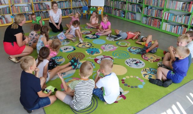dzieci i bibliotekarka siedzą na kolorowym dywanie, w tle regały z książkami
