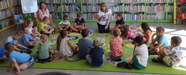 bibliotekarka z książką i przedszkolaki siedzą na kolorowym dywanie, w tle regały z książkami