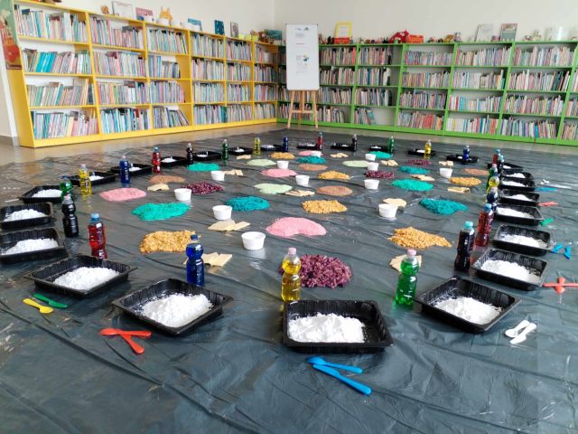 w sali bibliotecznej rozłożone na folii malarskiej w kółku tacki z różnymi materiałami, np. mąka, makaron, ryż