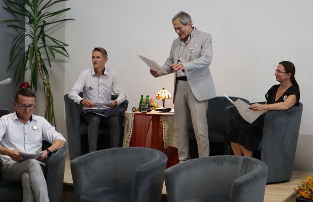 stojący aktor czytający tekst, na fotelach siedzący lektorzy