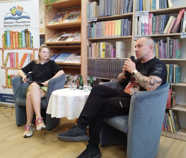 na zdjęciu autor z mikrofonem i prowadząca spotkanie, w tle regały z książkami