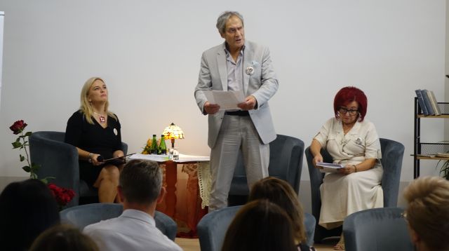 aktor stoi i czyta, w tle na fotelach siedzą pozostali czytający