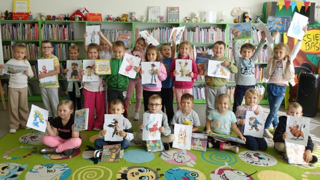 przedszkolaki pozują do zdjęcia z wykonanymi przez siebie pracami, w tle regały z książkami