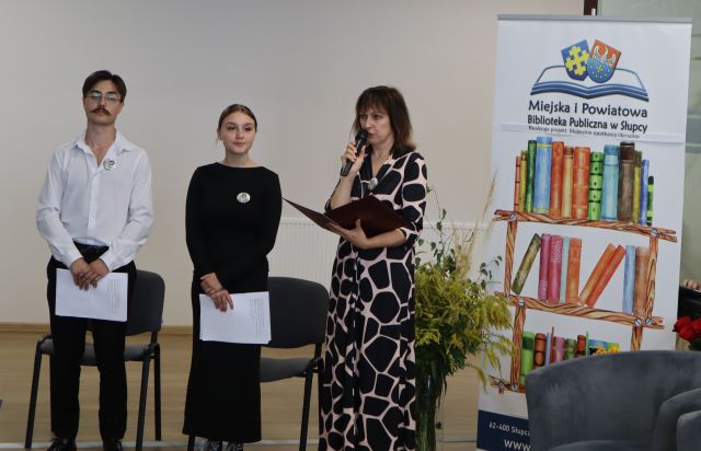 dyrektorka biblioteki wita zgromadzonych na uroczystości, obok stoi dziewczyna i chłopak prowadzący uroczystość