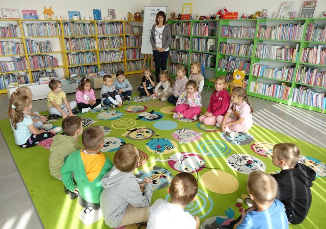 przdszkolaki siedzą na kolorowym dywanie, przed nimi dyrektorka biblioteki