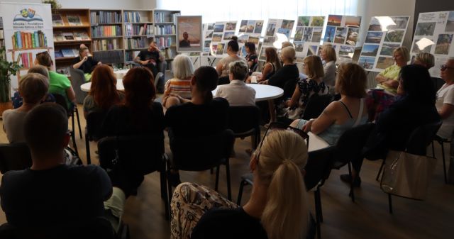 na zdjęciu uczestnicy spotkania siedzący na krzesłach i fotelach, przed nimi na fotelach siedzi autor książek i prowadząca spotkanie, w tle wystawa fotografii i regały z książkami