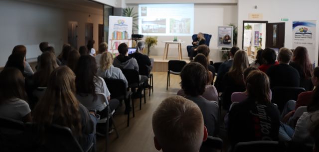 młodzież siedząca na krzesłach, w tle prezentacja wyświetlana na ekranie i siedząca na fotelu autorka książek