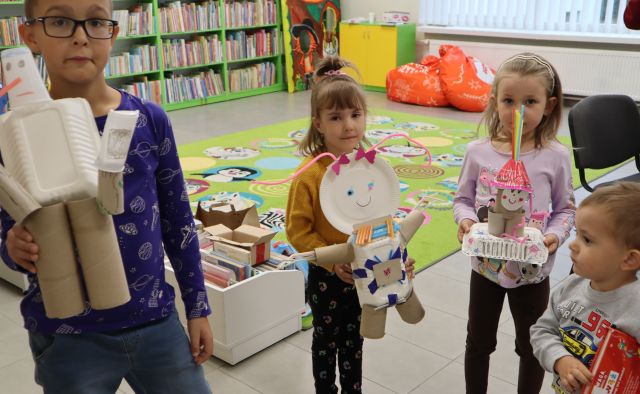5 dzieci prezentuje swoje roboty z materiałów recyklingowych, w tle regały z książkami