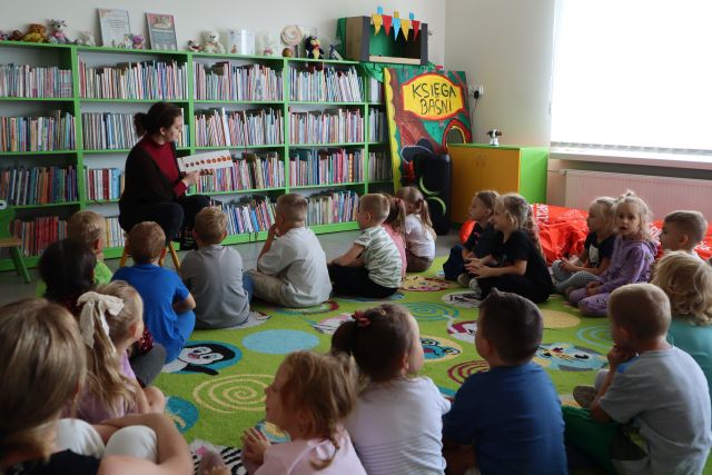 dzieci siedząc na kolorowym dywanie słuchają czytającej bibliotekarki, w tle regały z książkami