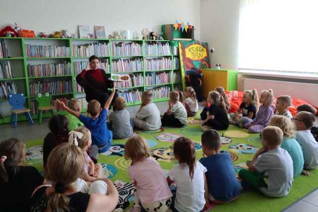 dzieci siedząc na kolorowym dywanie słuchają czytającej bibliotekarki, w tle regały z książkami