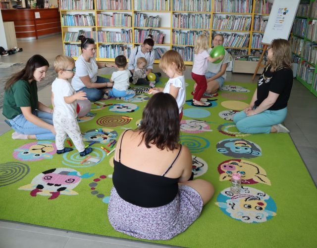 dzieci i rodzice bawią się balonami na kolorowym dywanie, w tle regały z książkami