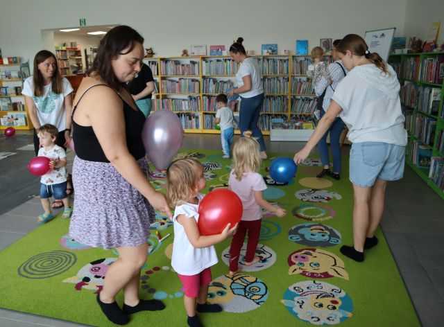 dzieci i rodzice bawią się balonami na kolorowym dywanie, w tle regały z książkami