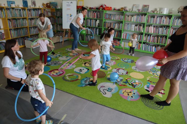 dzieci i rodzice bawią się balonami na kolorowym dywanie, zabawy z hula hop, w tle regały z książkami