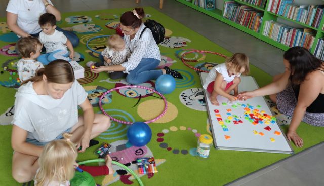 dzieci bawią się na kolorowym dywanie, siedzą przy nich rodzice