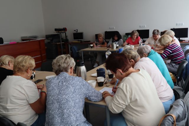 seniorki siedząc przy stołach rozwiązują zagadki logiczne