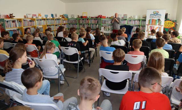 autor z mikrofonem stoi przed dziećmi uczestniczącymi w spotkaniu, w tle regały z książkami