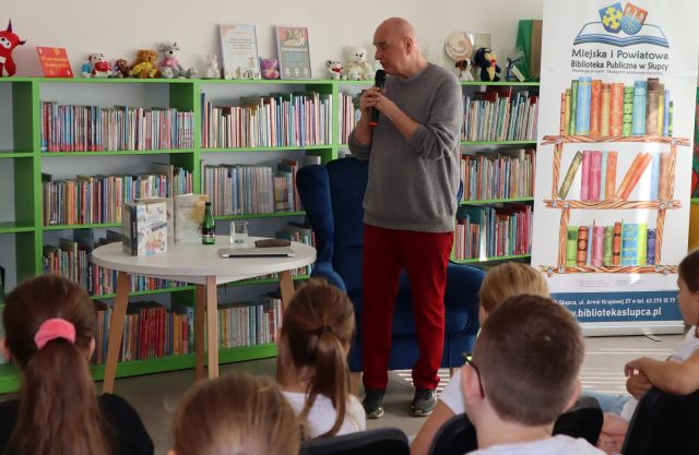 autor z mikrofonem mówi do siedzącej przed nim młodzieży, w tle regały z książkami