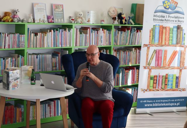 autor w okularach czyta dzieciom tekst z laptopa, w tle regały z książkami