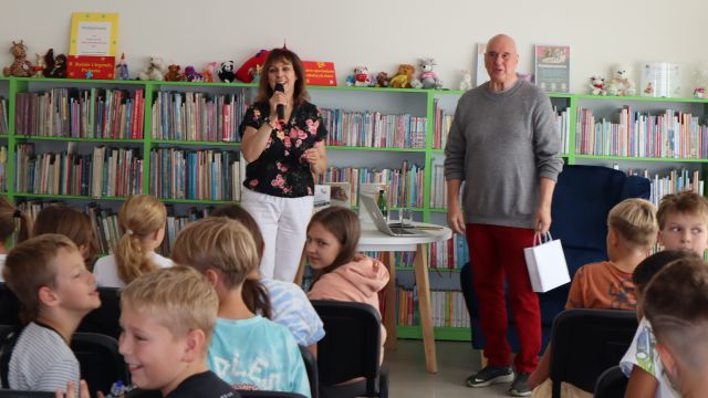dyrektorka biblioteki z mikrofonem, obok stoi autor, przed nimi siedzą na krzesłach dzieci, w tle regały z książkami