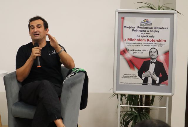 na zdjęciu aktor siedzący z mikrofonem, z prawej strony plakat reklamujący spotkanie