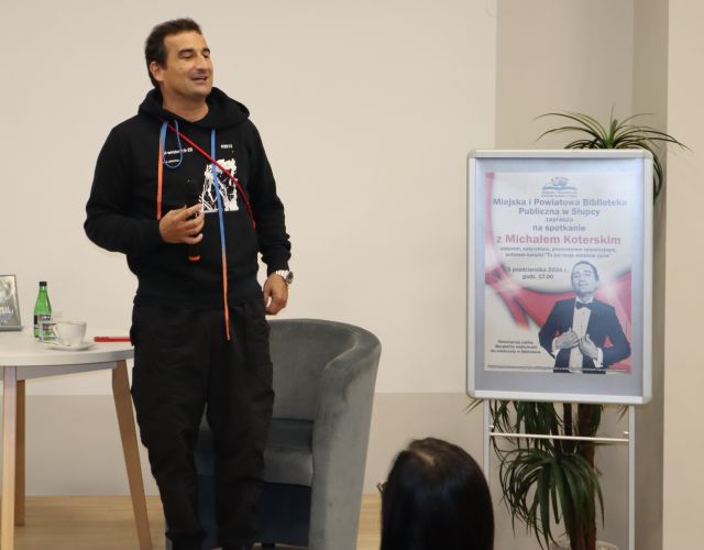 aktor stoi i przemawia przez mikrofon, z prawej strony plakat reklamujący spotkanie