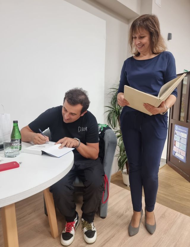 dyrektorka biblioteki stojąca z kroniką, aktor siedząc podpisuję książkę