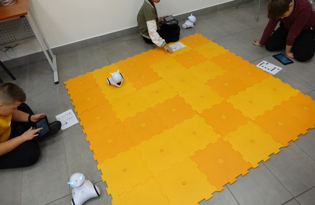 3 dzieci steruje robotami Photon jeżdżącymi po pomarańczowej macie przy użyciu tabletów