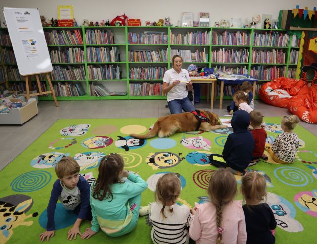kobieta z psem klęczy przed siedzącymi na dywanie dziećmi, w tle regały z książkami