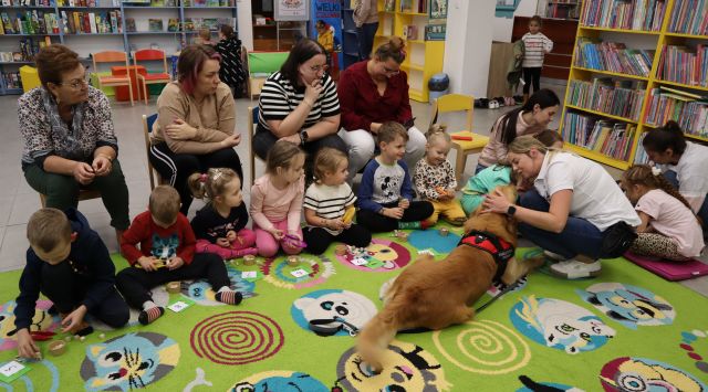 dzieci i pies siedzą na kolorowym dywanie, za dziećmi na krzesłach siedzą rodzice