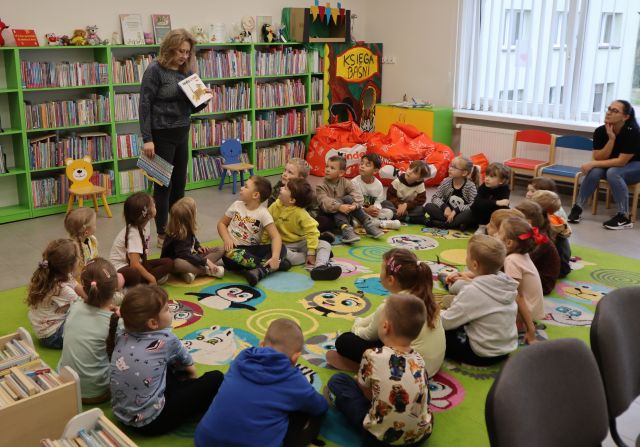 bibliotekarka opowiada dzieciom siedzącym na dywanie, w tle regały z książkami