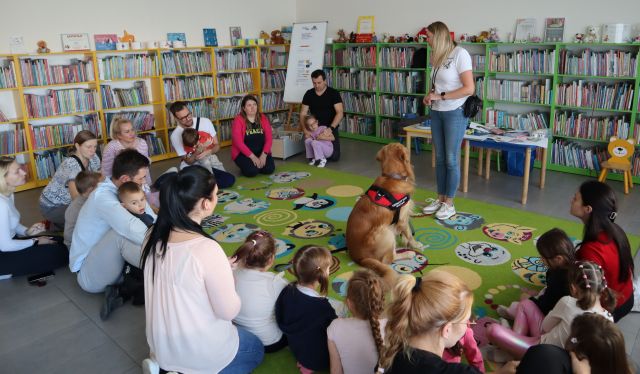 dzieci z rodzicami siedzą na kolorowym dywanie w kółku, przed nimi stoi kobieta, a w środku siedzi pies