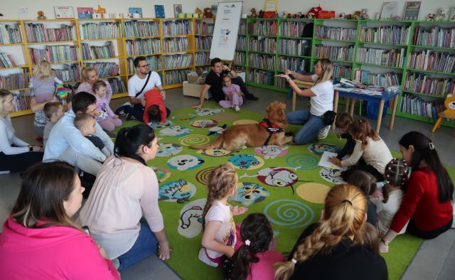 dzieci z rodzicami siedzą na kolorowym dywanie w kółku, przed nimi kuca kobieta, a w środku siedzi pies
