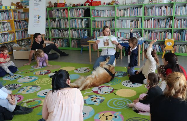 kobieta pokazuje dzieciom zdjęcia z psem, przed nią siedzi pies i dzieci z rodzicami w okręgu
