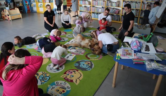na środku dywanu leży pies na plecach, wokół siedzą dzieci i rodzice