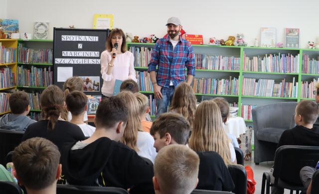dyrektorka biblioteki z mikrofonem i pisarz stoją przed siedzącą młodzieżą, w tle regały z książkami