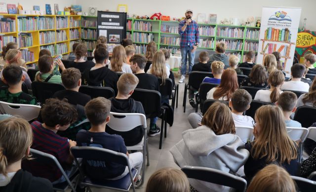 młodzież siedzi na krzesłach, przed nimi stoi autor, w tle regały z książkami