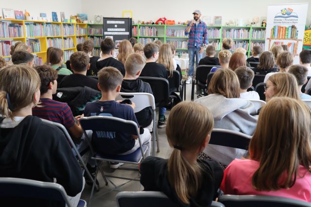 młodzież siedzi na krzesłach, przed nimi stoi autor z mikrofonem, w tle regały z książkami