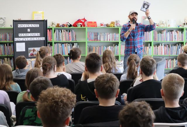 autor z mikrofonem stoi przed siedzącą na krzesłach młodzieżą, w tle zielone regały z książkami