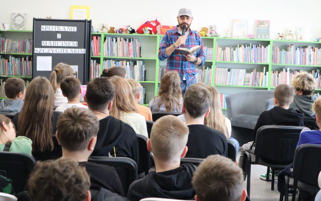 autor z mikrofonem czyta dzieciom siedzącym na krzesłach książkę, w tle regały z książkami