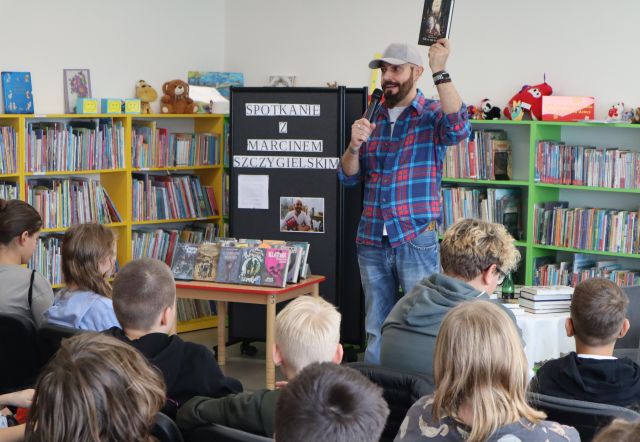 autor z mikrofonem pokazuje młodzieży siedzącej na krzesłach książkę, w tle regały z książkami