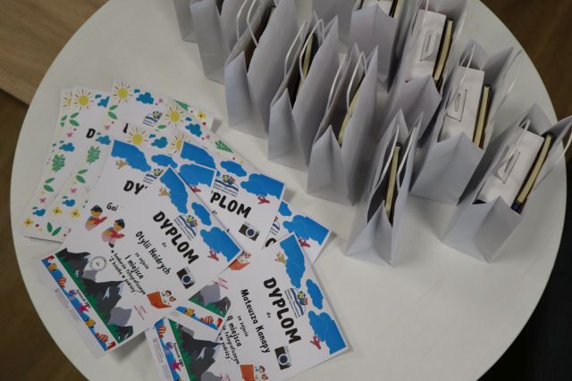 na białym okrągłym stole rozłożone dyplomy dla laureatów oraz nagrody w białych torebkach