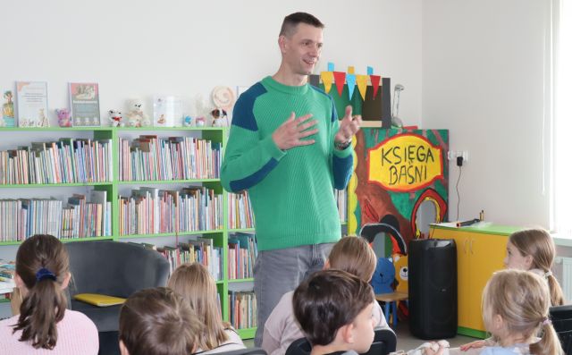 autor w zielonej bluzie stoi przed siedzącą na krzesłach młodzieżą, w tle regały z książkami