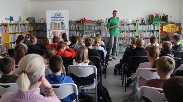 autor w zielonej bluzie stoi przed siedzącą na krzesłach młodzieżą, w tle regały z książkami